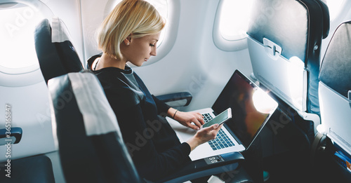 Businesswoman with short haircut sitting in airplane cabin and chatting online on smartphone while checking email on laptop computer with mock up area.Female traveler reading notification on cellular