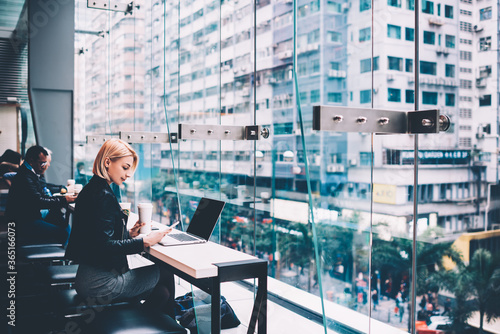 Beautiful blonde young woman with coffee in hand reading received notification on smartphone while working freelance at modern laptop computer with internet and mock up area in cafe with cityspace photo