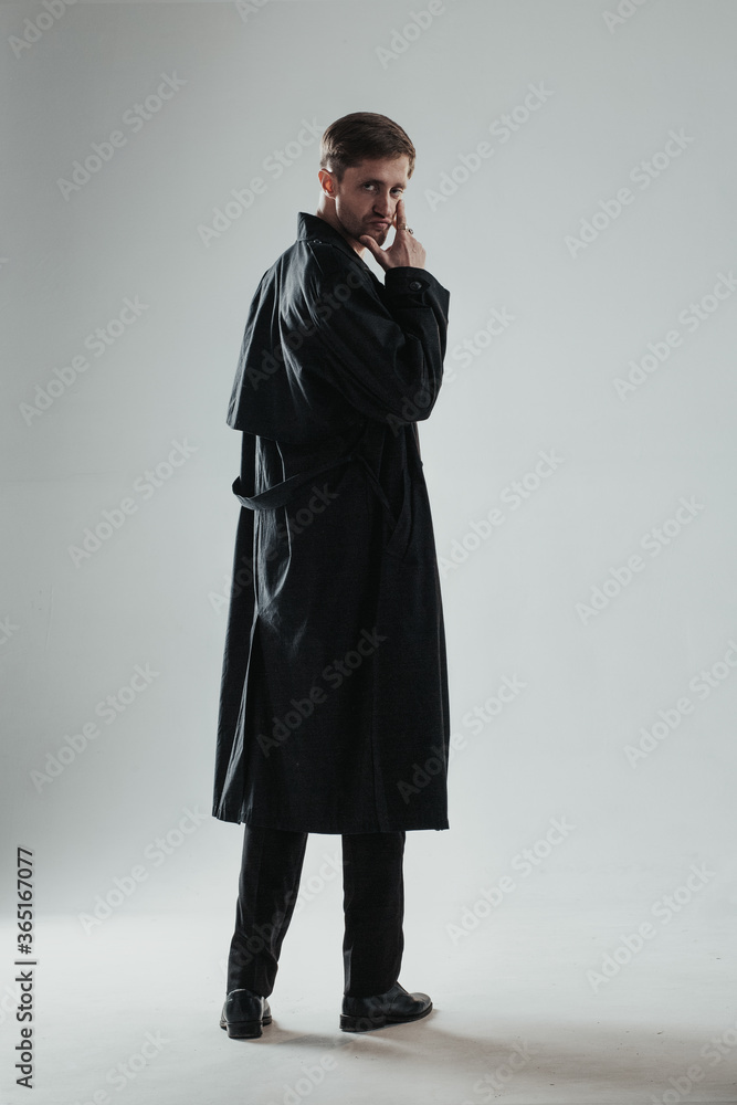 Young and handsome caucasian man posing in a black trench coat in the studio with gray background. Vertical image. Young and handsome caucasian man posing in a black trench coat in the studio with gray background. Vertical image.