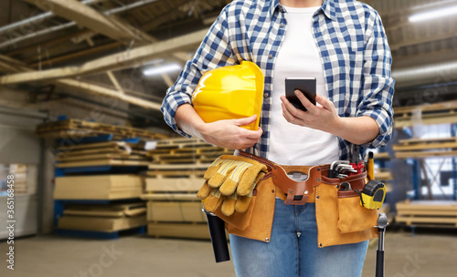 industry, production and job concept - close up of woman or worker with smartphone, helmet and working tools on belt over factory workshop on background © Syda Productions