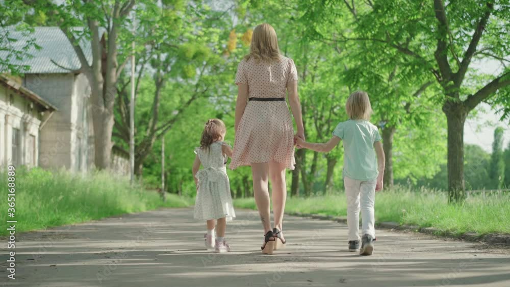 Back view of young blond Caucasian mother walking with two children along the alley in park. Wide shot of happy Caucasian family strolling outdoors on sunny summer day. Lifestyle, unity.