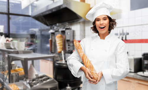 cooking, culinary and bakery concept - happy smiling female chef or baker in toque holding french bread or baguette over restaurant or kebab shop kitchen background