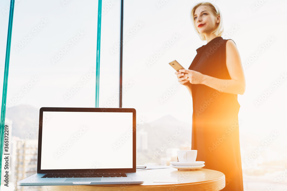 Confident female owner of a successful company is holding mobile phone in hands, while is standing in modern office interior near table with open portable net-book with mock up copy space screen Confident female owner of a successful company is holding mobile phone in hands, while is standing in modern office interior near table with open portable net-book with mock up copy space screen