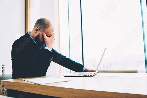 Tired male financial employee depressed about failed business project sitting at laptop computer connected to wireless high speed internet.Stressful entrepreneur in formal wear fatigued from hard work