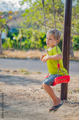 Sad lonely boy sit on swing look far away, wait for friends or while parents are busy, thinking about important things for him. Summer, childhood, leisure, friendship, relationship and people concept