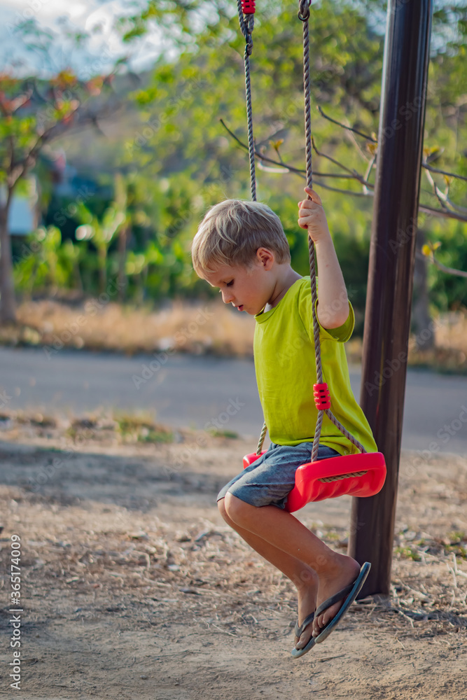 Sad lonely boy sitting on swing looking down on ground, waiting for friends or while parents are busy. Summer, childhood, leisure, friendship and people concept Sad lonely boy sitting on swing looking down on ground, waiting for friends or while parents are busy. Summer, childhood, leisure, friendship and people concept