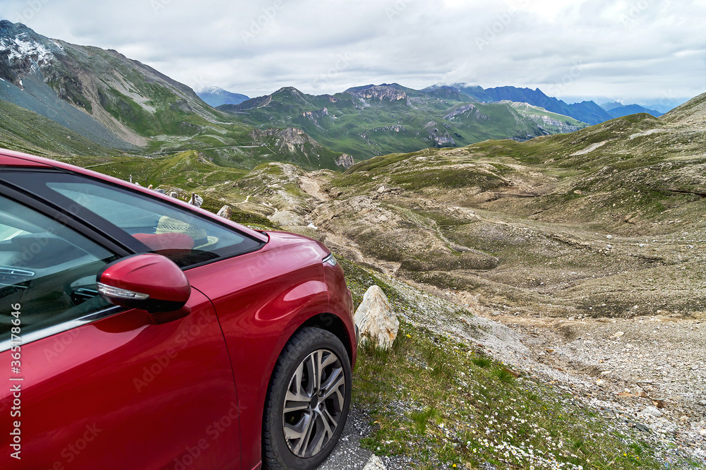 Naklejka premium Red car for traveling is standing on the mountain road in the Alps, Austria. Amazing landscape with snowy mountain peaks and sky on background. Family vacation, travel, transportation concept.