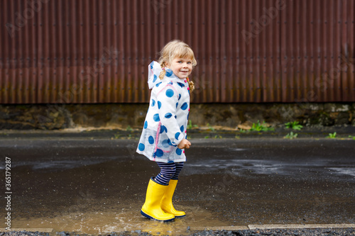 Little toddler girl wearing yellow rain boots, running and walking during sleet on rainy cloudy day. Cute happy child in colorful clothes jumping into puddle, splashing with water, outdoor activity