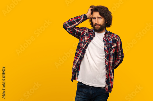 A man in a plaid shirt reflects and holds and scratches his head, looks to the side on a yellow background
