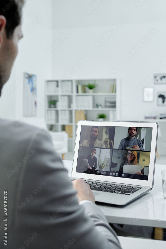 Over shoulder view of businessman sitting at desk in office and talking to colleagues via video conferencing on laptop