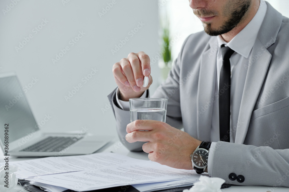 Close-up of bearded businessman sitting at desk with papers and laptop and taking hangover prevention pill in office Close-up of bearded businessman sitting at desk with papers and laptop and taking hangover prevention pill in office