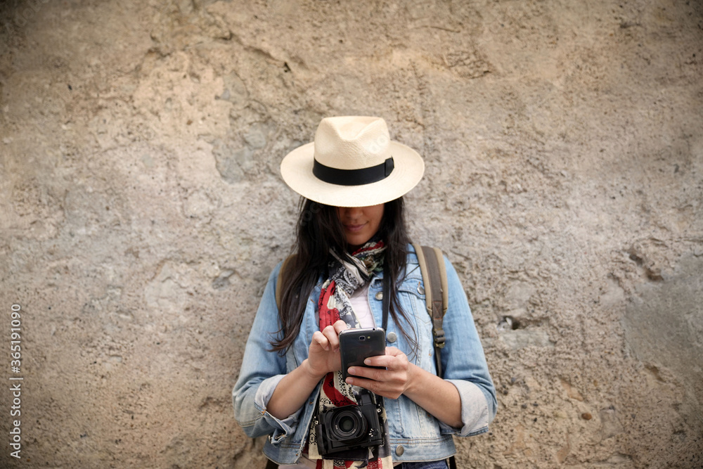 Woman wearing blue jeans and hat, standing against wall and using smartphone