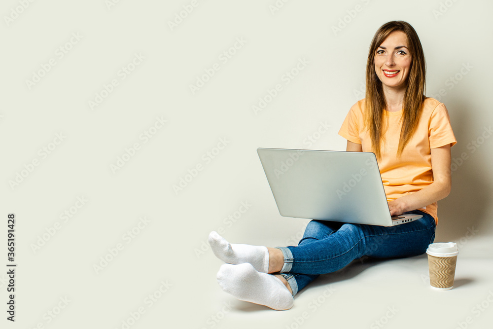 Naklejka premium Smiling young woman holding a laptop on her knees, a paper cup with coffee is standing nearby on a light background
