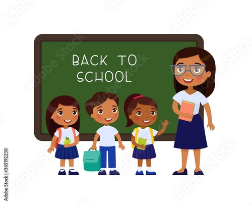 Teacher greeting pupils in classroom flat vector illustration. Boys and girls dressed in school uniform and female teacher pointing at blackboard cartoon characters. Primary students back to school