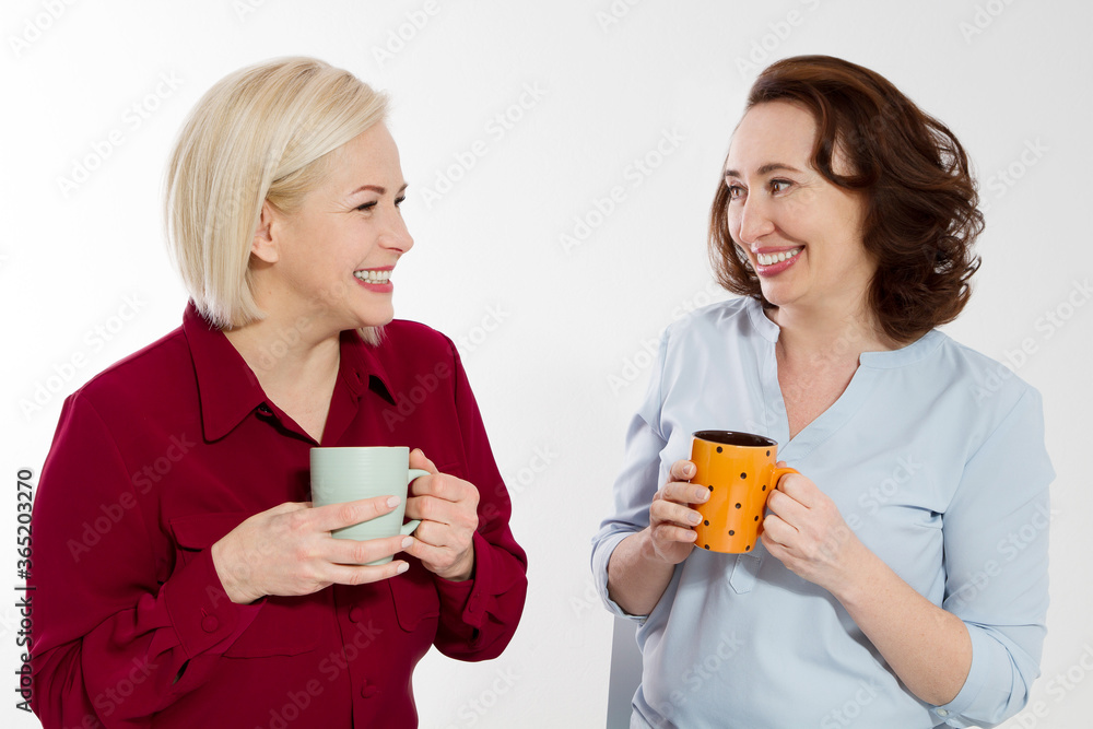Two healthy and beautiful middle aged women drinking tea and talking isolated on white background. Woman support woman. Happy menopause concept. Two healthy and beautiful middle aged women drinking tea and talking isolated on white background. Woman support woman. Happy menopause concept.