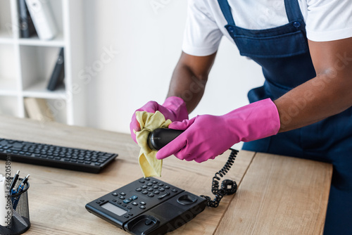 Cropped view of african american cleaner using rag while cleaning telephone on office table