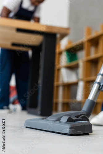 Selective focus of brush of vacuum cleaner on floor and african american cleaner in office