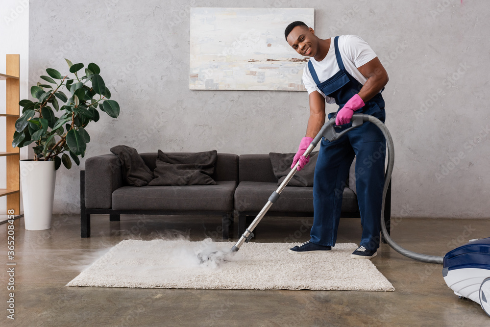 Fototapeta premium Smiling african american cleaner in overalls using vacuum cleaner with hot steam at home
