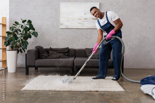Smiling african american cleaner in overalls using vacuum cleaner with hot steam at home