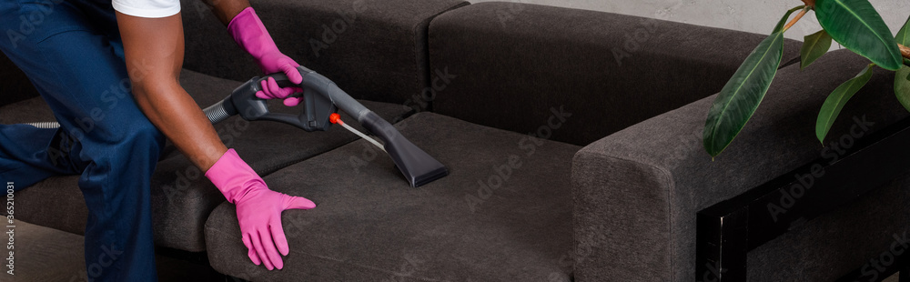 Panoramic shot of african american cleaner cleaning couch with vacuum cleaner at home