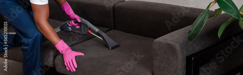 Panoramic shot of african american cleaner cleaning couch with vacuum cleaner at home