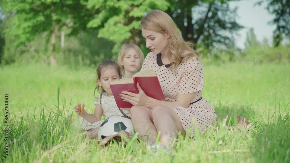 Blond Caucasian woman with tattoo on leg reading book on green meadow as little boy and girl sitting at back. Portrait of positive mother spending summer weekend with son and daughter. Blond Caucasian woman with tattoo on leg reading book on green meadow as little boy and girl sitting at back. Portrait of positive mother spending summer weekend with son and daughter.