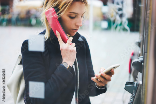 Pensive young female traveler dialing number of international code browsing information via 4g connection on modern smartphone while calling using telephone booth standing outdoors on city street photo