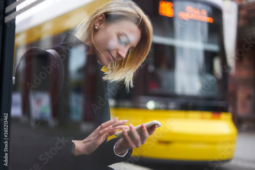 Smiling blonde hipster girl waiting for public transport on bus stop chatting in social networks via smartphone,casually dressed female tourist using application for searching traffic schedule