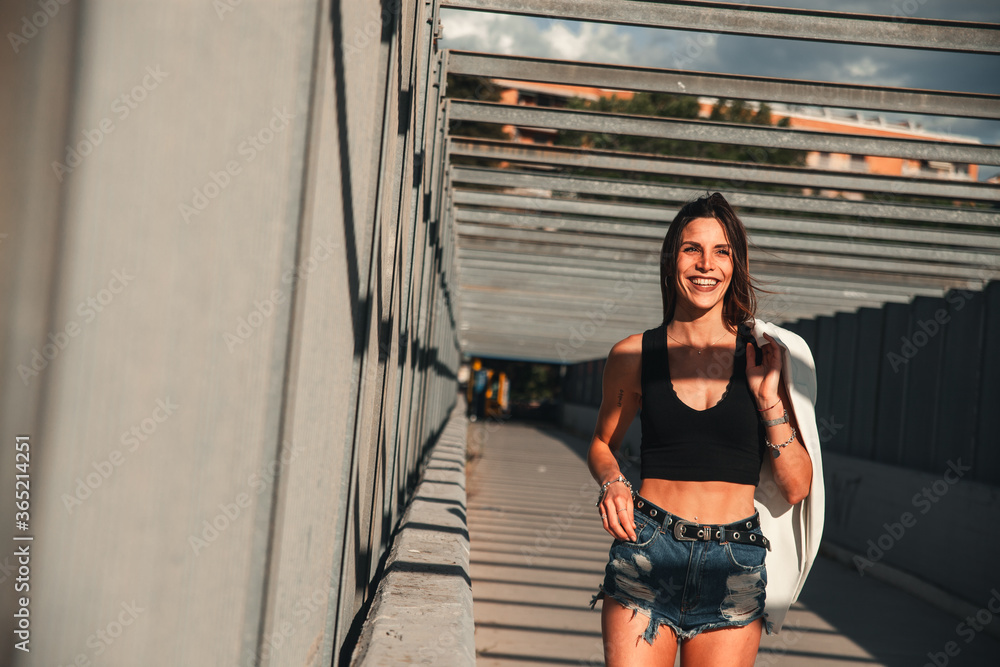 Scatto in prospettiva in una località metropolitana di una modella affascinante che cammina sorridente indossando un top nero e con in mano una giacca bianca Scatto in prospettiva in una località metropolitana di una modella affascinante che cammina sorridente indossando un top nero e con in mano una giacca bianca
