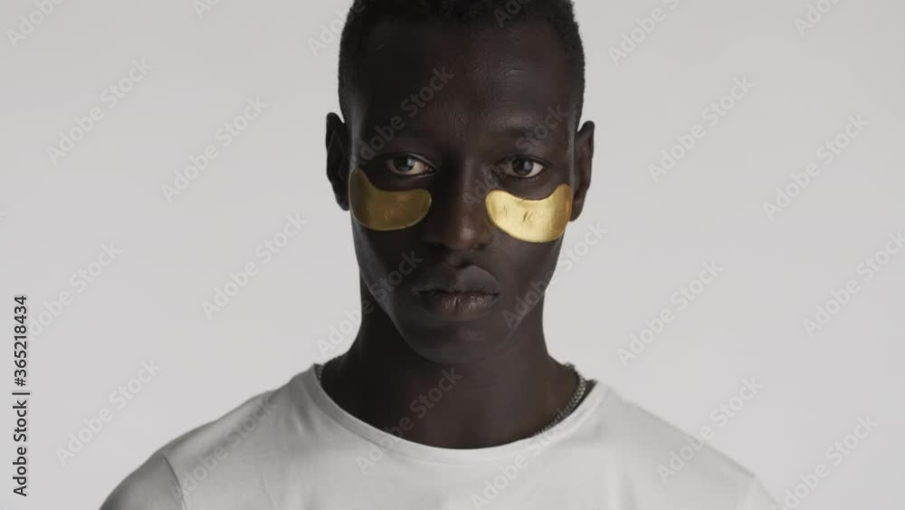 Portrait of handsome serious African American man with gold eye patches intently looking in camera isolated. Facial treatment Portrait of handsome serious African American man with gold eye patches intently looking in camera isolated. Facial treatment