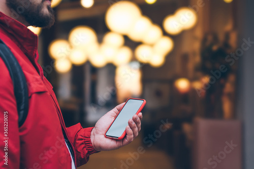 Side cropped view of male person in red jacket holding modern cellphone device with blank mock up screen outdoors at evening street. Men's hands keeps mobile phone near copy space with bokeh lights