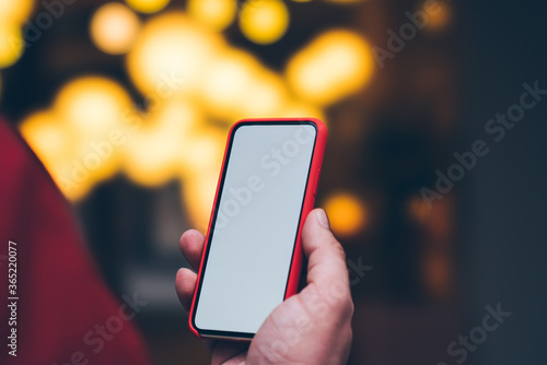 Side close up view of male person holding modern smartphone with copy space screen while standing outdoors near bokeh lights background. Man using mobile phone applications with internet connection