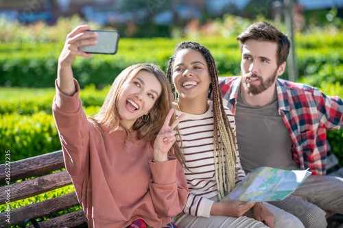Girl with smartphone taking selfie with friends on bench.