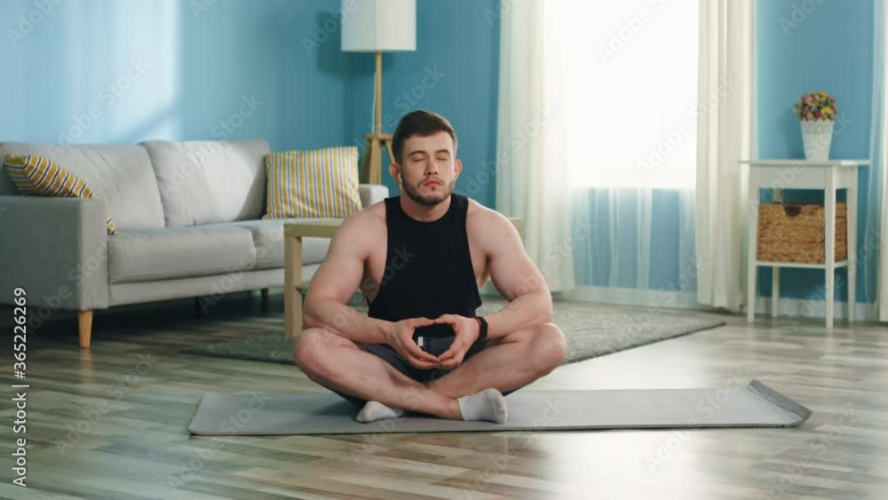 Young athletic man is sitting on a floor at home and practices calm breathing, trying to relax and to recall good memories, listen favorite music, enjoys new day, Slow motion. Young athletic man is sitting on a floor at home and practices calm breathing, trying to relax and to recall good memories, listen favorite music, enjoys new day, Slow motion.