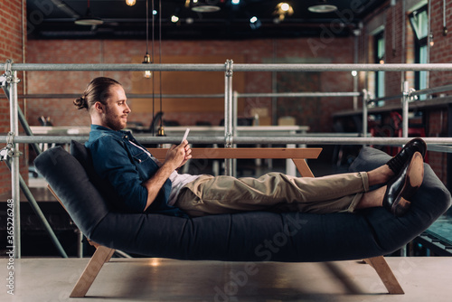 side view of businessman chilling on sofa and using smartphone in office