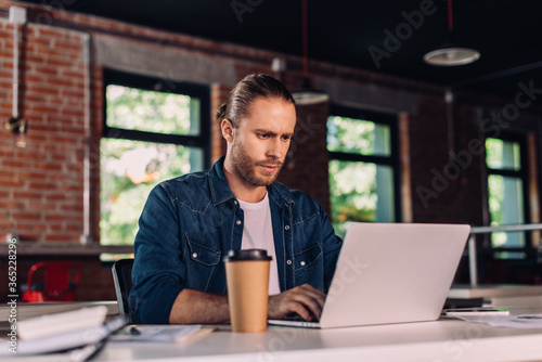 selective focus of businessman looking at laptop near paper cup on table