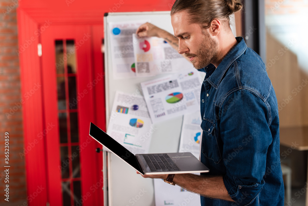 selective focus of handsome businessman looking at laptop near charts and graphs selective focus of handsome businessman looking at laptop near charts and graphs