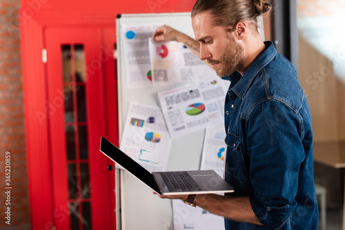 selective focus of handsome businessman looking at laptop near charts and graphs