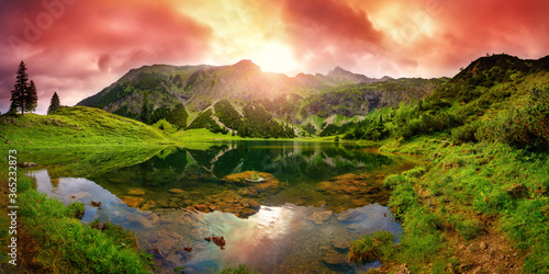 Dramatic sunrise at a lake in the Alps with mountains, red clouds reflected in the clear water and paths leading through the vibrant green grass