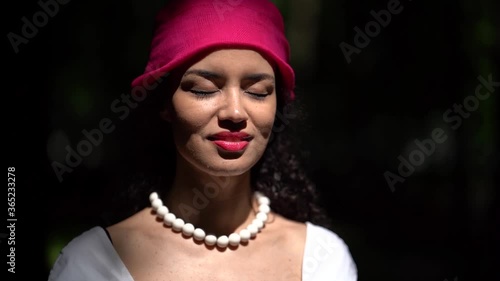 Close-up of a dark-haired curly-haired young smiling girl walking in a green summer Park. She wears large white beads and a lilac headscarf. She stands with her eyes closed and then opens them.