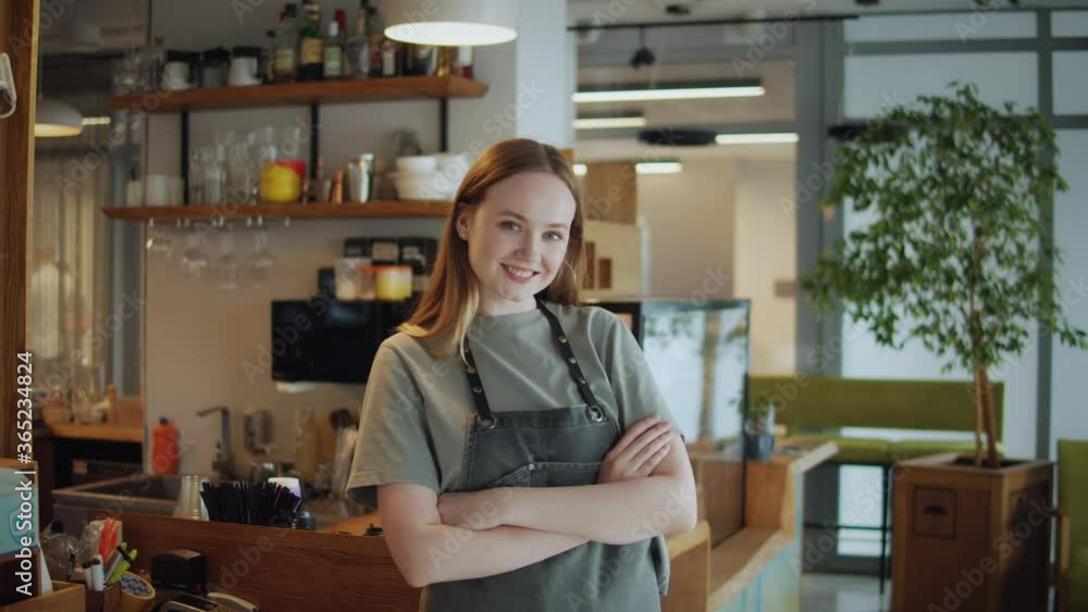 Portrait of happy woman standing in her cafe coffee shop. Cheerful young waitress waiting for clients at coffee shop. Successful small business owner in casual wearing grey apron standing at entrance Portrait of happy woman standing in her cafe coffee shop. Cheerful young waitress waiting for clients at coffee shop. Successful small business owner in casual wearing grey apron standing at entrance