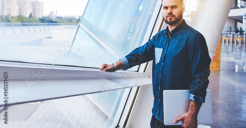 Confident man with serious face holding closed net-book in hand while standing near office window indoors, successful male freelancer posing in modern interior after presentation on laptop computer