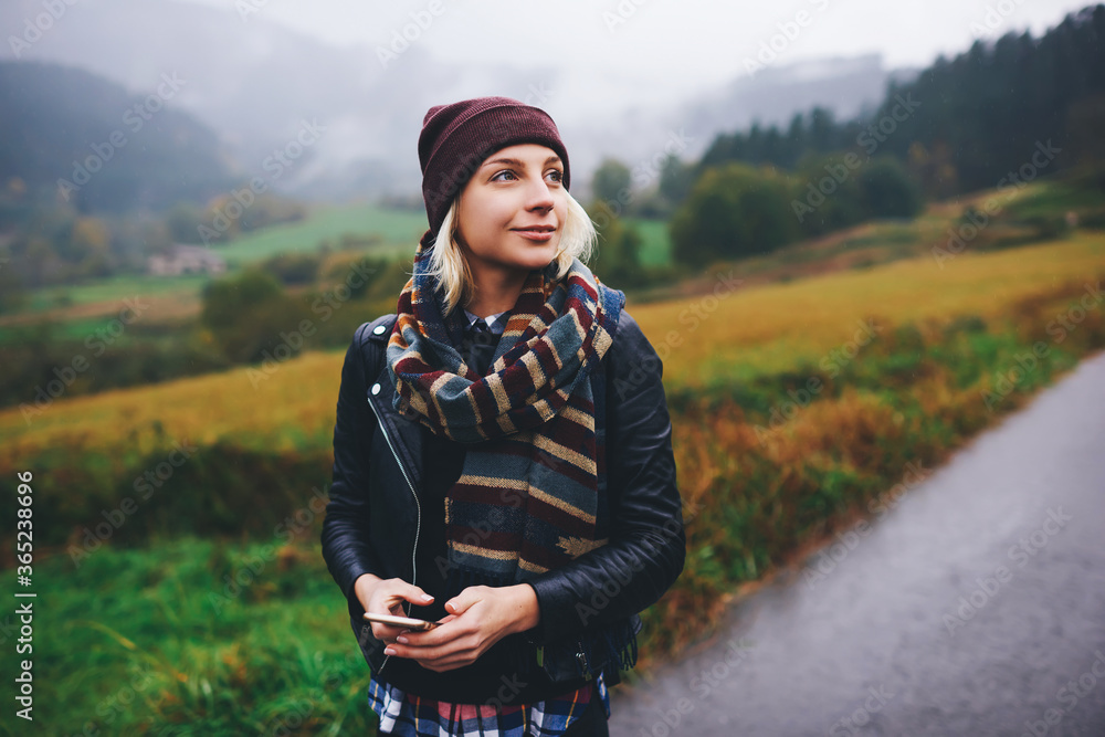 Young female wrapped up in warm scarf using electronic map on cell telephone while hiking on a mountain trail, charming woman tourist holding mobile phone while admires amazing nature view in fall day