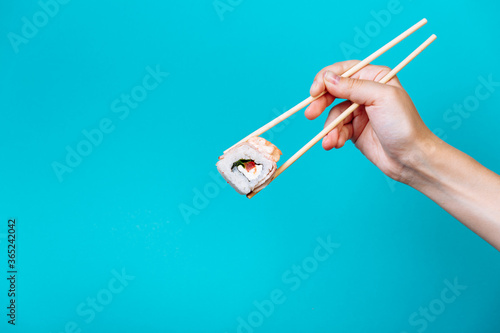 Woman's hand holds Tasty sushi roll maki with wooden chopsticks on blue background close up. Place for caption and text