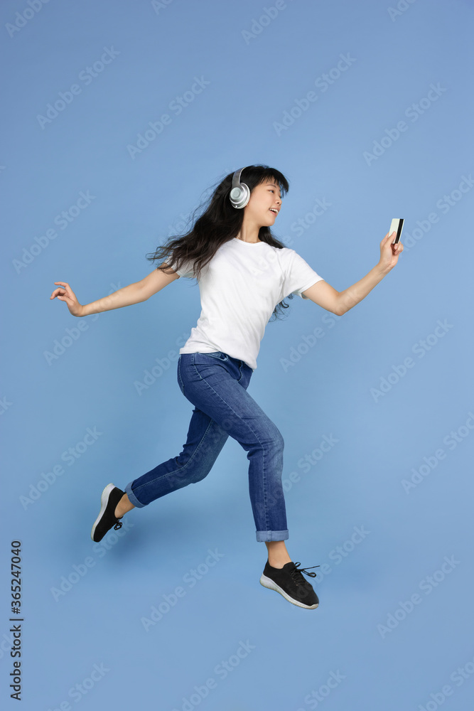 Jumping high with card and smartphone. Portrait of young asian woman isolated on blue background. Beautiful cute girl. Human emotions, facial expression, sales, ad, online shopping concept.