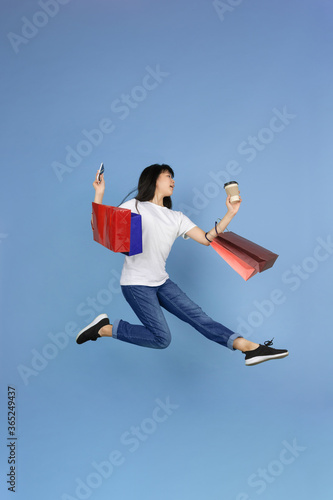 Jumping high with shopping bags and phone, coffee. Portrait of young asian woman isolated on blue background. Beautiful cute girl. Human emotions, facial expression, sales, ad, online shopping concept