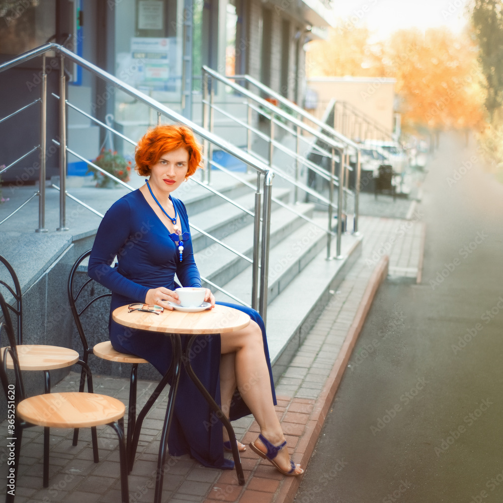 Beautiful looking red haired 30 years old woman sitting in cafe with cup of coffee. Time for yourself in summer day. Morning tea or coffee in modern street restaurant Beautiful looking red haired 30 years old woman sitting in cafe with cup of coffee. Time for yourself in summer day. Morning tea or coffee in modern street restaurant