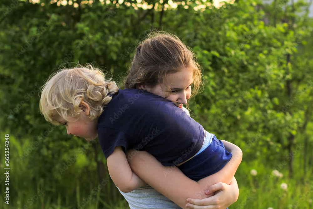 Fototapeta premium Cute teenager girl and little blonde boy playing and laughing on a green meadow. Sister and brother, siblings. Summertime, family concept. 