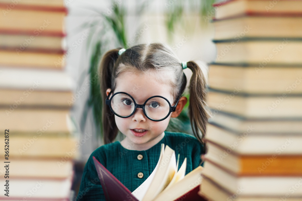 Cute smiling preschool age girl in round big glasses with red book in her hands between two books piles Cute smiling preschool age girl in round big glasses with red book in her hands between two books piles