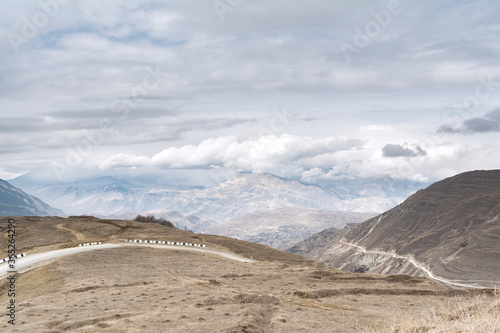 View of the alpine road against the background of the mountain peaks of a gloomy sky with clouds and dry grass of sand color. The concept of landscape, travel.